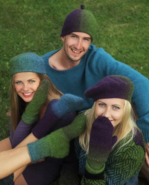 Tre unge mennesker som sitter tett sammen på en gressplen og viser frem luer, votter og sokker